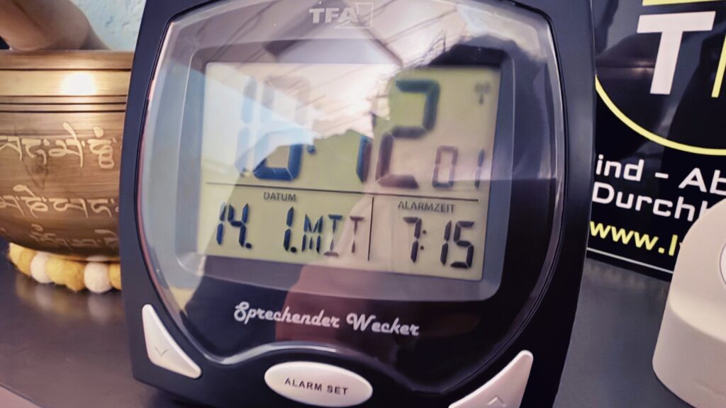Das Bild zeigt eine vergrößerte Ansicht des Weckers, der auf einem Regal steht. Im Bild sind außerdem eine Klangschale und ein Schild mit der Aufschrift "Blind - Aber mit Durchblick" zu sehen. Der Wecker zeigt 18:12 Uhr an am 14. 1. Die Alarmzeit ist auf 7:15 Uhr gestellt. Unter der Anzeige befindet sich der Text "Sprechender Wecker", darunter auf dem Knopf steht "Alarm Set".