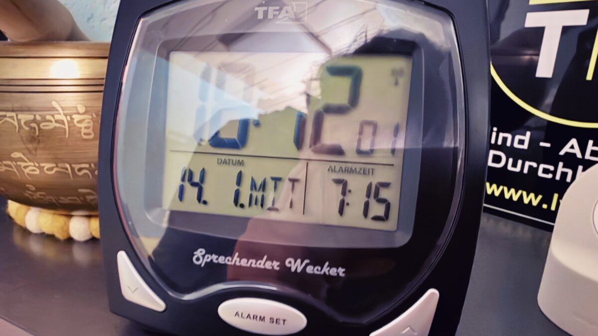 Das Bild zeigt eine vergrößerte Ansicht des Weckers, der auf einem Regal steht. Im Bild sind außerdem eine Klangschale und ein Schild mit der Aufschrift "Blind - Aber mit Durchblick" zu sehen. Der Wecker zeigt 18:12 Uhr an am 14. 1. Die Alarmzeit ist auf 7:15 Uhr gestellt. Unter der Anzeige befindet sich der Text "Sprechender Wecker", darunter auf dem Knopf steht "Alarm Set".