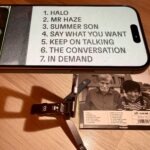 Das Bild zeigt ein iPhone 15, welches auf dem Stativ montiert ist. Das Stativ steht auf der CD "The Muscle Shoals Sessions" von Texas. Vergrößert wird die Tracklist.