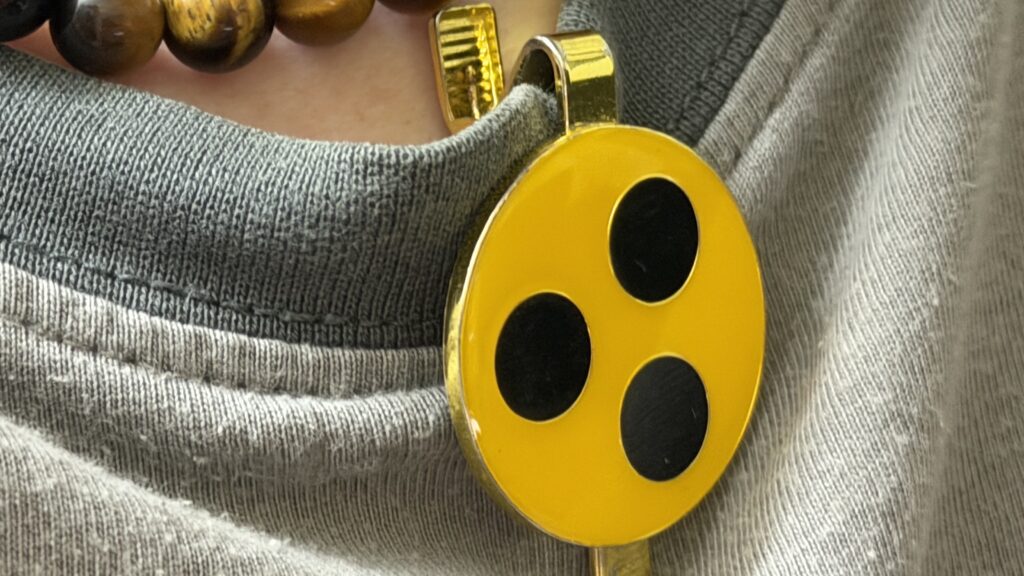 Das Bild zeig ein gelbes Blindenabzeichen mit 3 schwarzen Punkten als Button zum anklemmen. Dieser befindet sich an einem T-Shirt-Kragen.