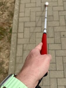 Ausgefalteter Blindenstock, gehalten in der linken Hand, nach vorne gestreckt, rollt auf dem Gehweg.